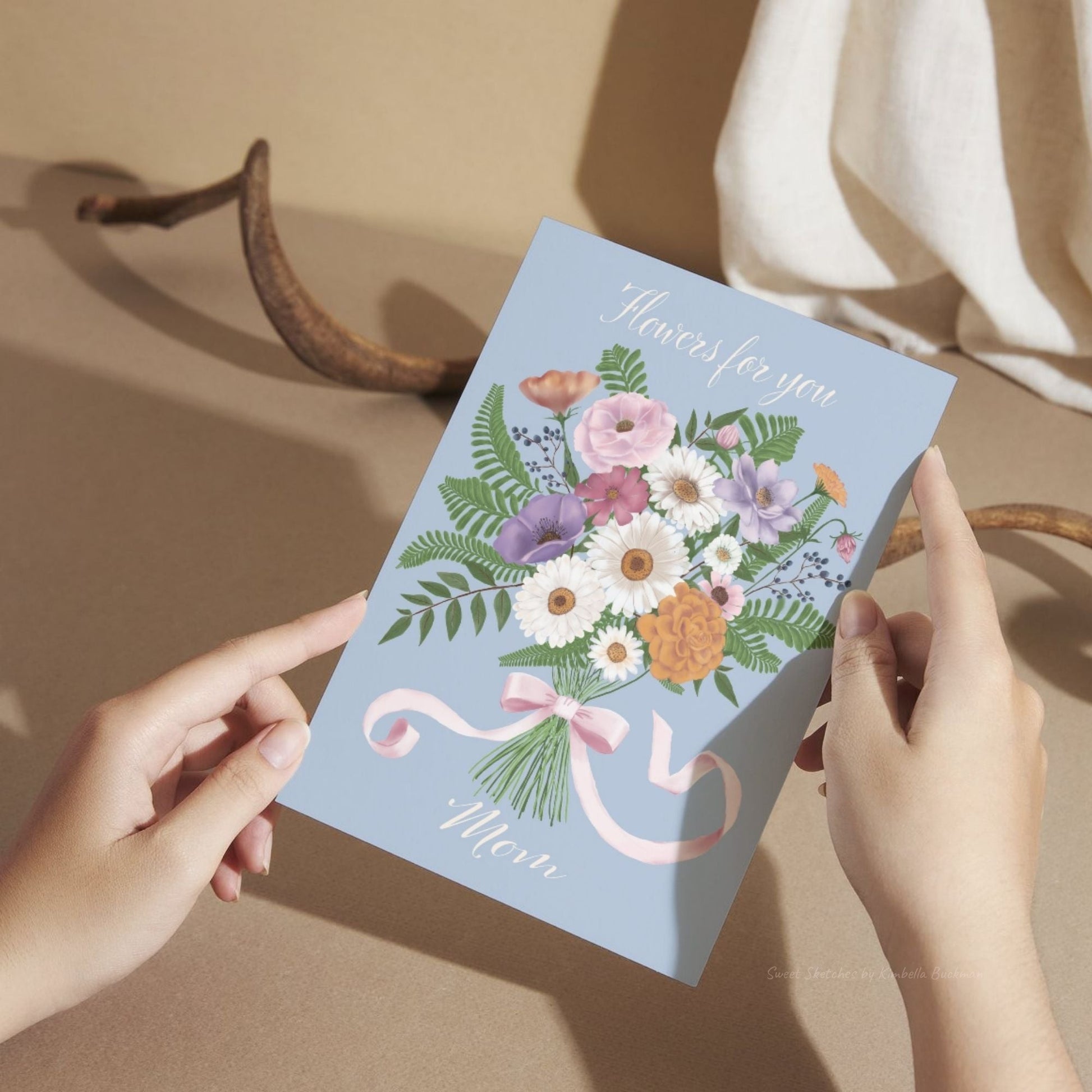 Hands holding a floral Mother's Day greeting card with a blue background and bouquet tied with a pink ribbon that reads “Flowers for you Mom.”